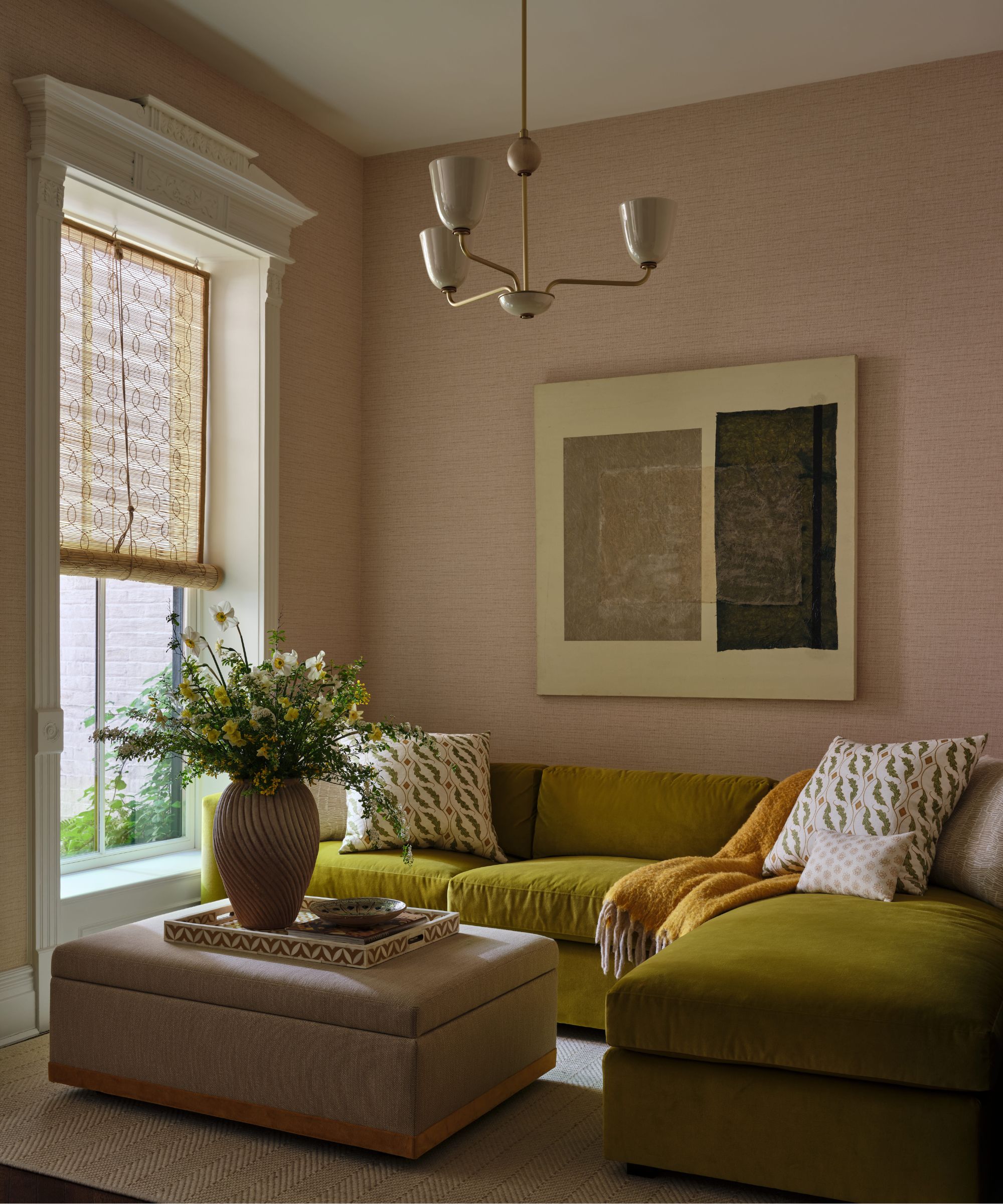 a bijoux family room space with grasscloth wallcovering and a large green velvet sectional couch, ottoman and window with chik blind