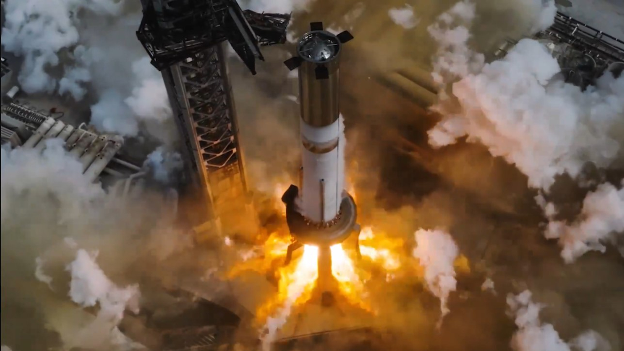 A tall chrome rocket booster stands next to a launch tower as huge explosions of fire burst from its engines below. Smoke billows in all directions. SpaceX's Super Heavy booster ignites its 33 Raptor engines during a static test fire July 15, 2024.