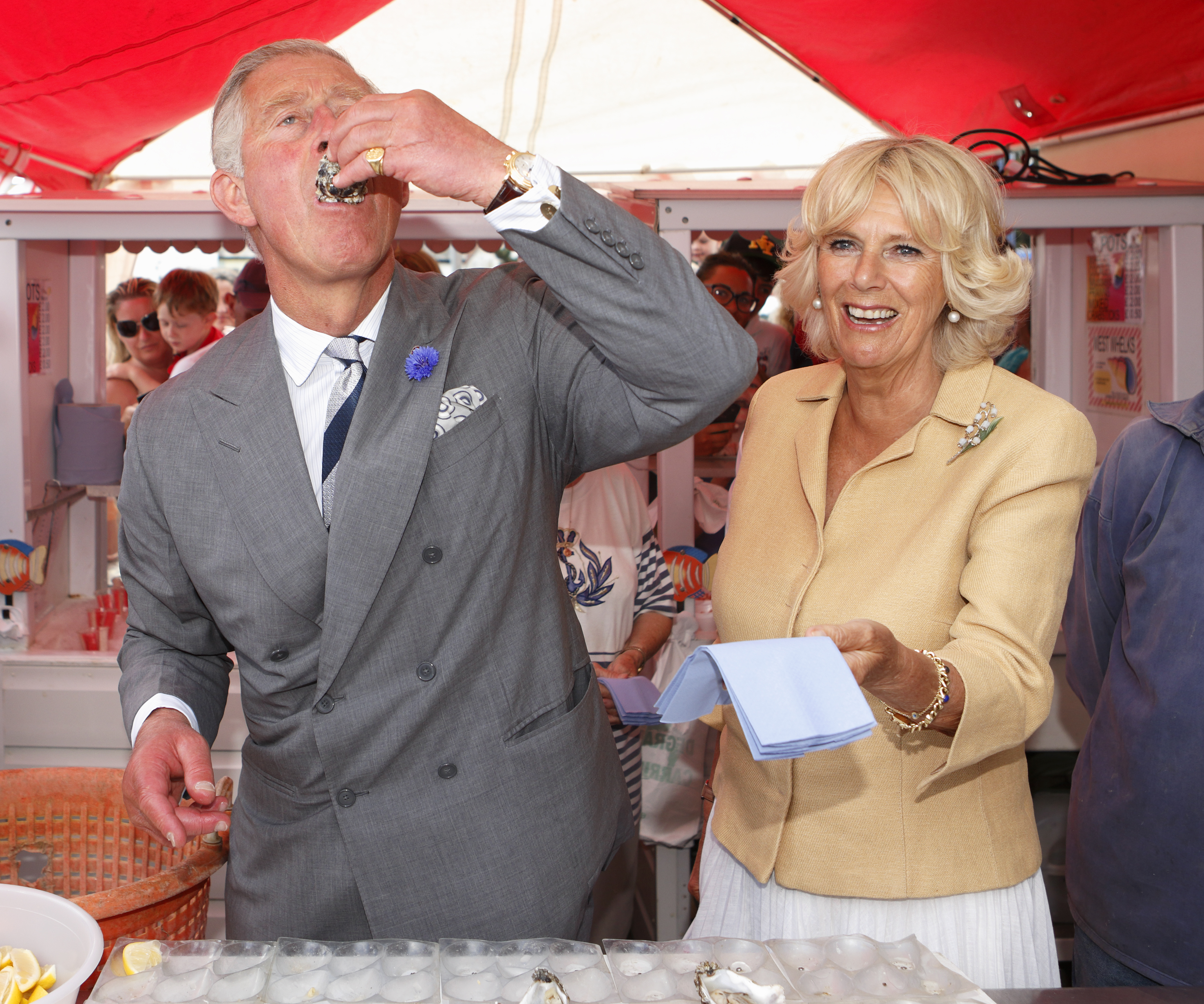 King Charles eating an oyster while Queen Camilla laughs next to him and holds a napkin