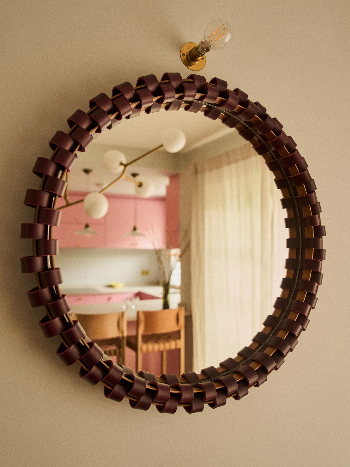 A leather-clad, round mirror reflecting a pink kitchen.