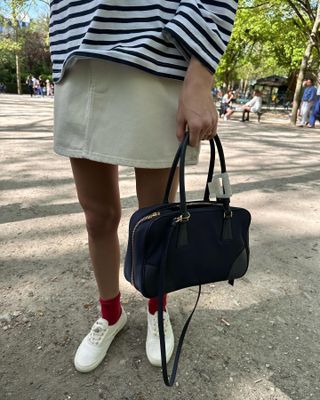 Waist-down photo of a woman wearing khaki skirt, striped long-sleeve shirt, Prada bag, and white sneakers with red socks.