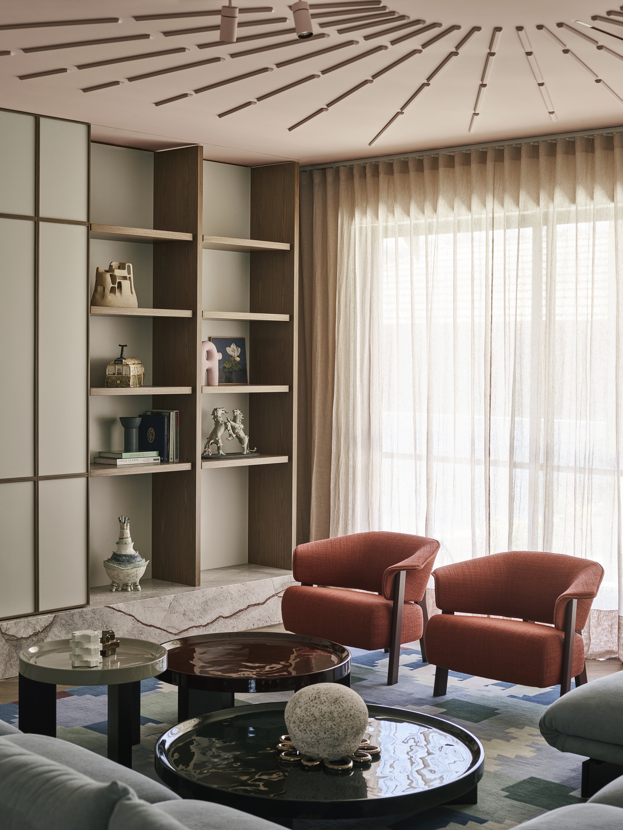modern living room with pink ceiling with decorative detail, cabinetry, two red armchairs, patterned rug, nesting coffee tables, and blue sofa