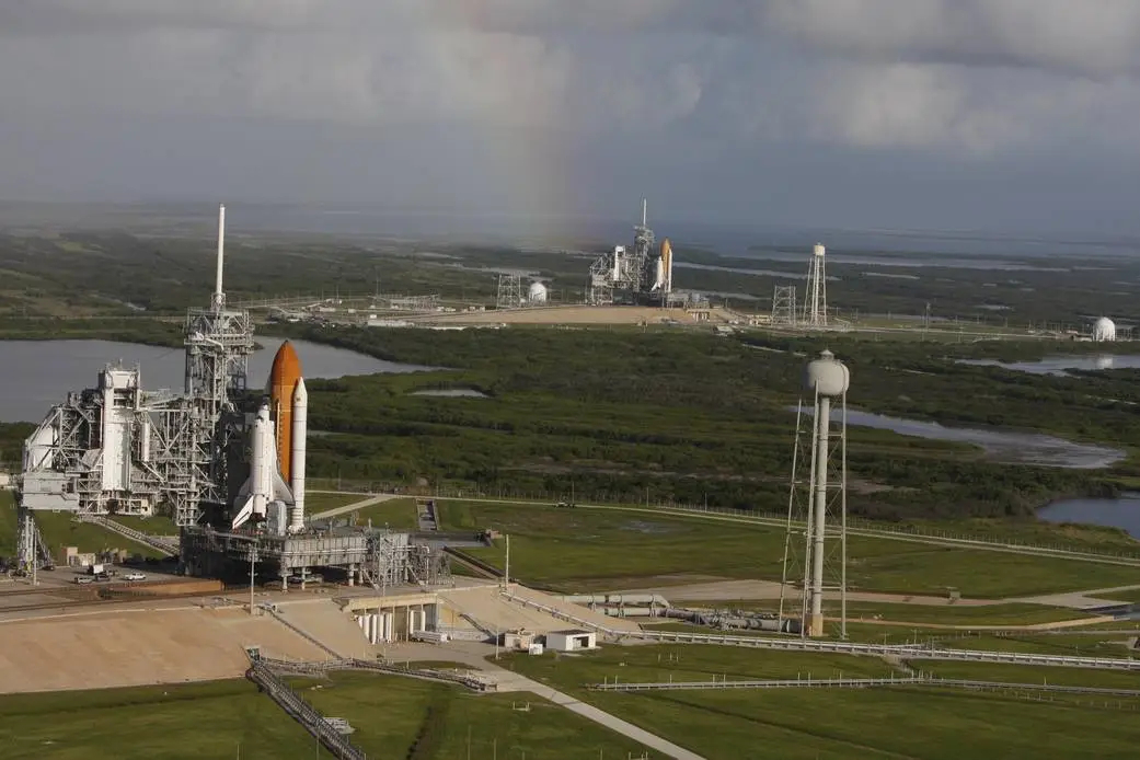 a space shuttle stands on the launchpad, seen from high, with another space shuttle standing on a different launchpad in the distance. A rainbow shines. NASA space shuttle Atlantis (foreground) at the agency's Kennedy Space Center in Florida, being readied for its STS-125 Hubble Space Telescope mission with backup shuttle Endeavour (far in background) also on the launch pad. After Atlantis returned safely, Endeavour was re-tasked for the STS-126 mission to the International Space Station.
