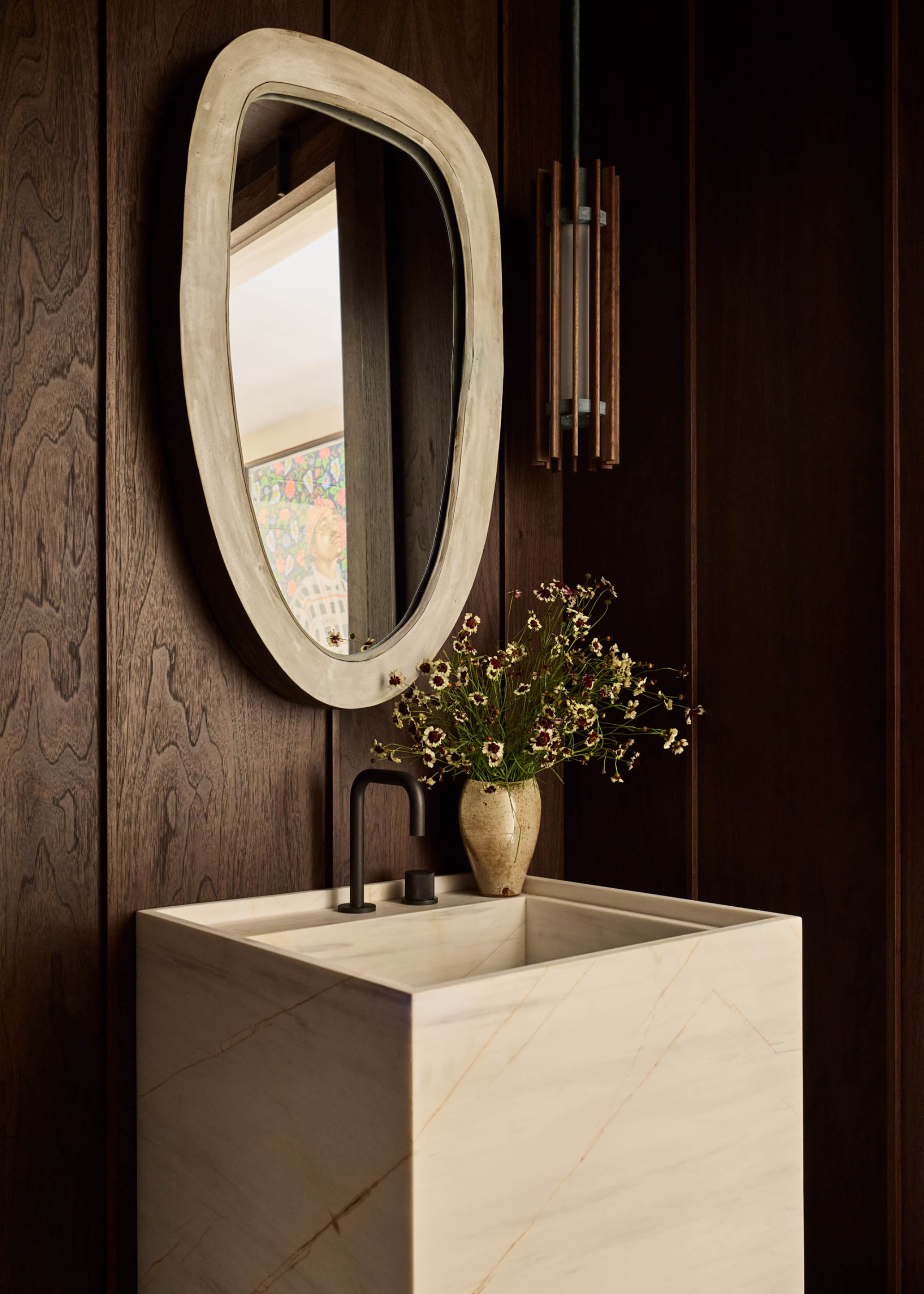 A dark wood powder room with an oval mirror, a marble sink with a vase of flowers and a pendant lamp