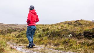 A woman walks along a muddy path, wearing a bright red jacket and Salewa Mountain Trainer 2 Mid Gore-Tex Boots.