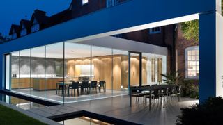 luxurious glass box kitchen extension with frameless glazing windows, two dining tables both outdoor and indoor, and a modern kitchen inside with wooden cabinets and island