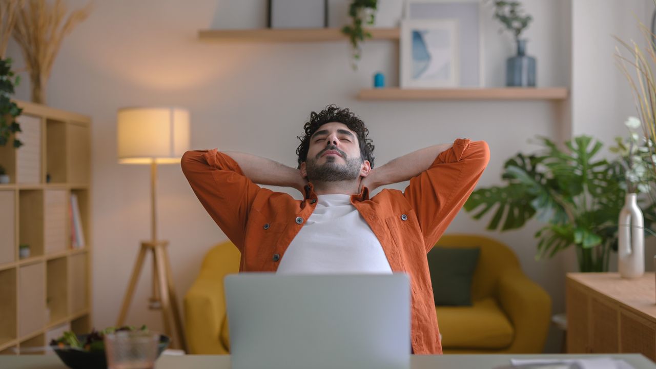 A man in a t-shirt and shirt stretches at a desk. He is facing the camera, with hands clasped behind his head and eyes closed. He leans back slightly in his chair. Behind him we see a sofa, a lamp, shelving and leafy plants.