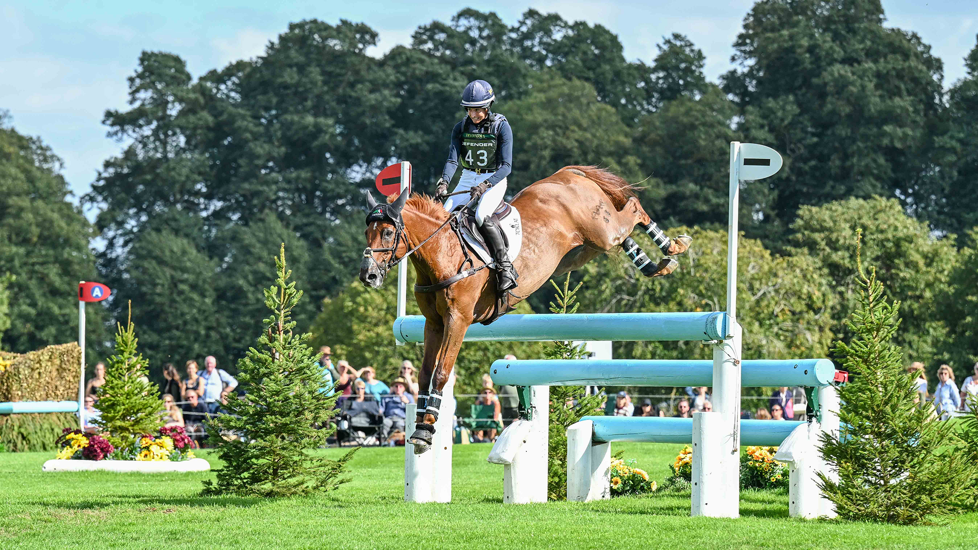 Jennie Brannigan, a rider in a blue top and hat cover, rides chestnut horse FE Lifestyle over a turquoise triple bar on the Burghley cross-country course
