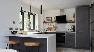 Contemporary kitchen with a minimalist aesthetic, featuring black pendant lights, a grey countertop, and wooden bar stools. 