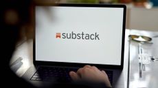 Substack logo and branding pictured on a laptop screen with hand placed on keyboard and blurred user head in foreground.