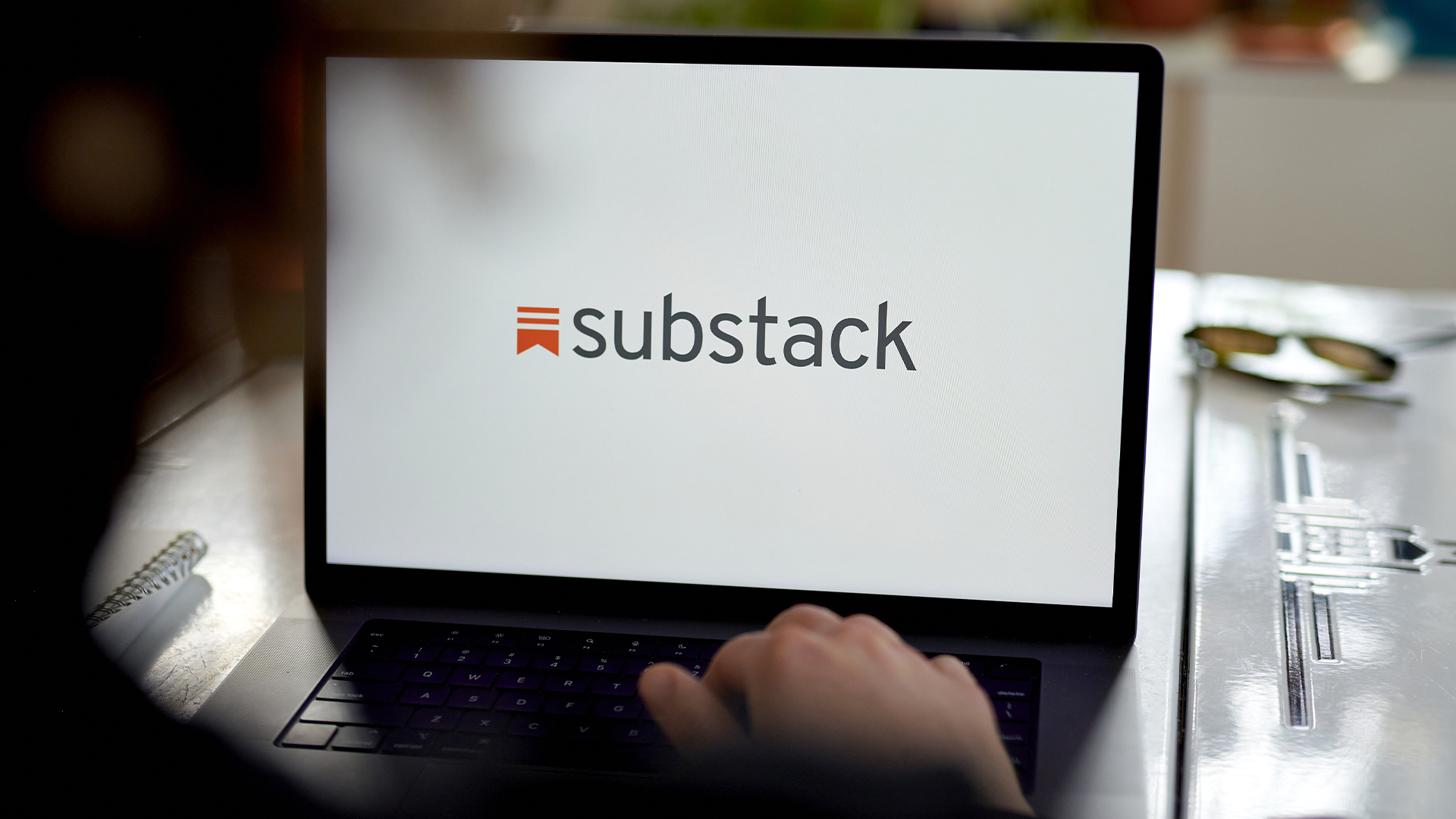 Substack logo and branding pictured on a laptop screen with hand placed on keyboard and blurred user head in foreground.