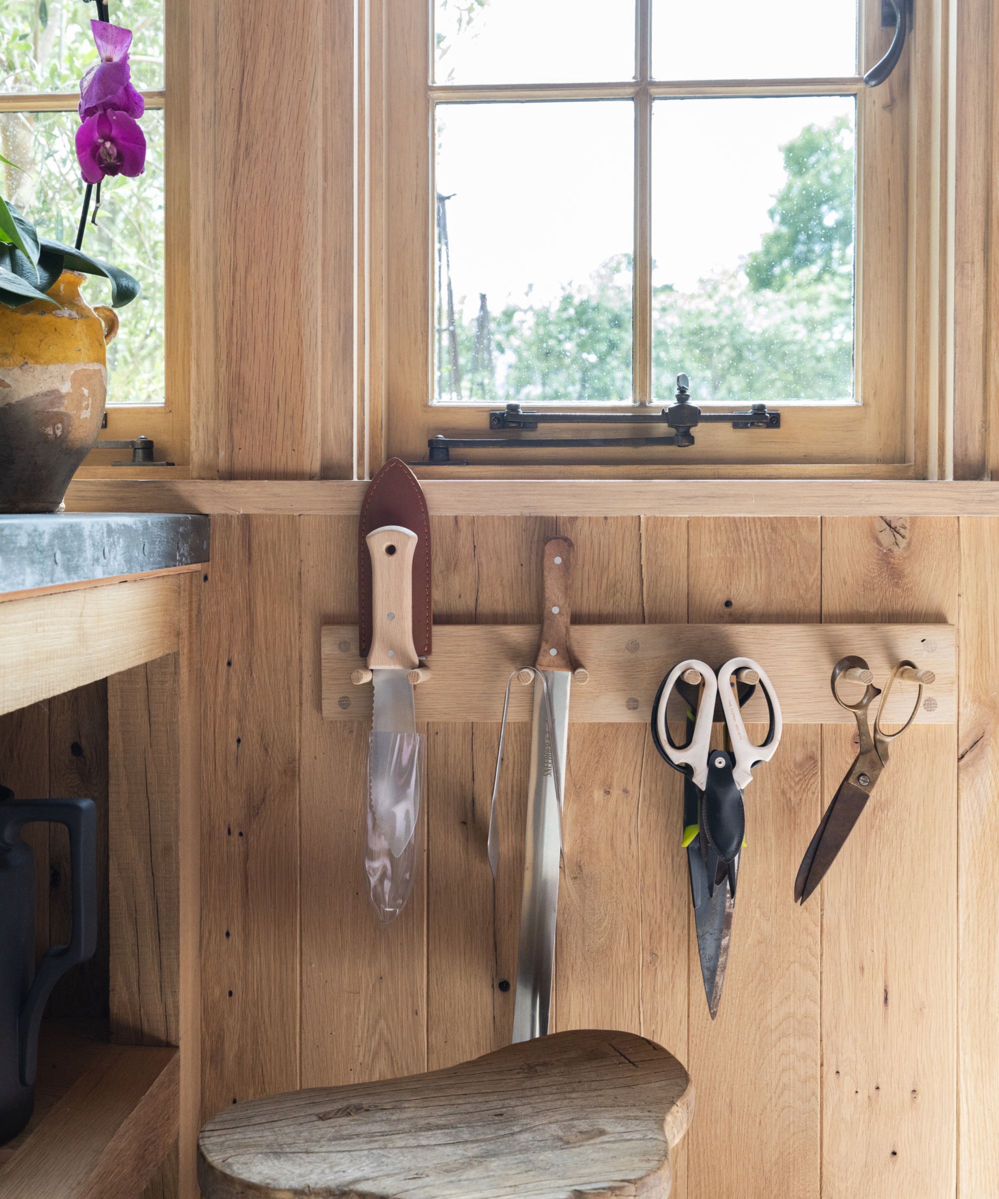A close-up of a small wooden peg rack on a wooden shed wall, with trowels, knives, and scissors hanging from it.