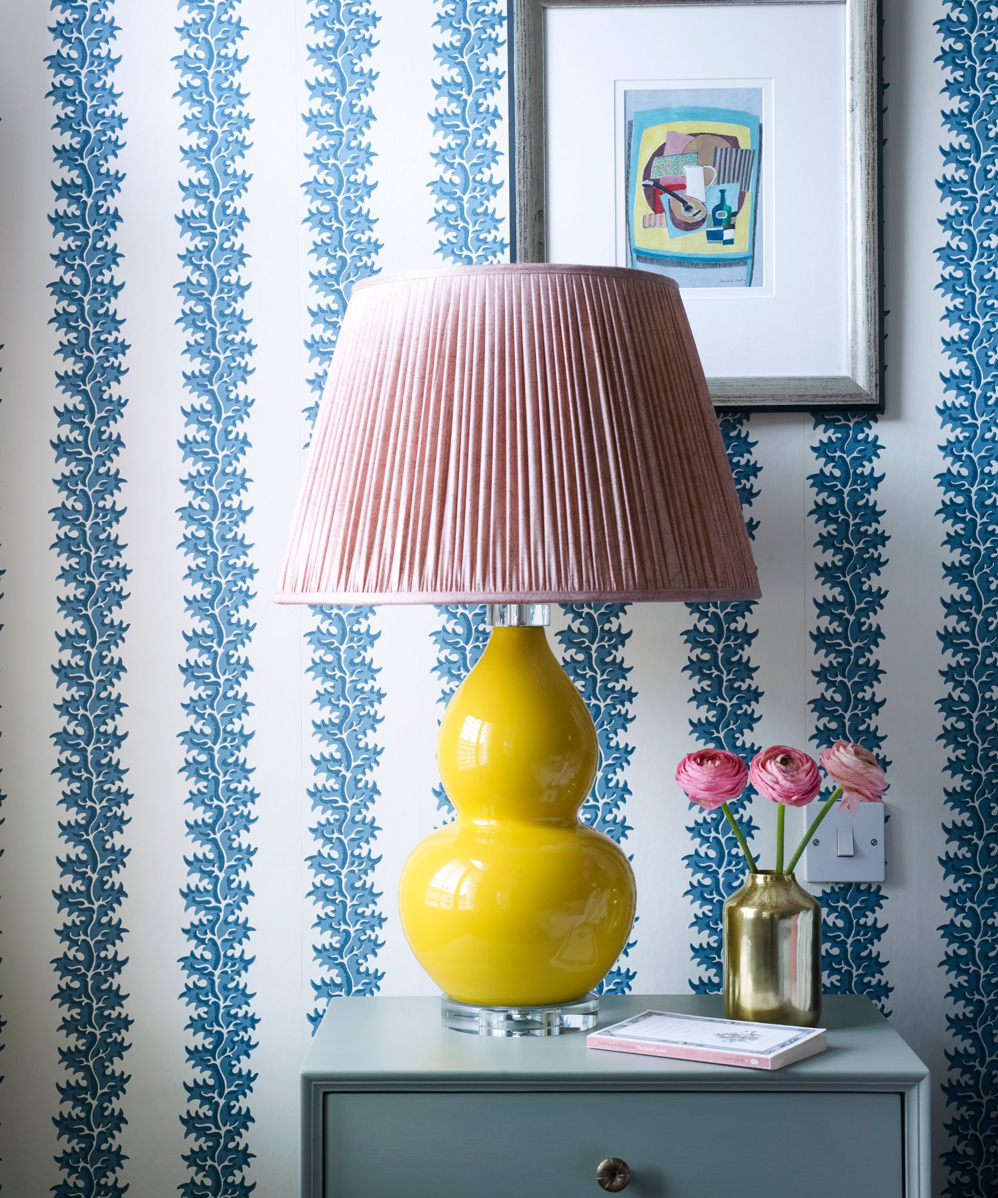 A room with blue and white statement wallpaper, a yellow ceramic lamp with pink lampshade, framed artwork in silver frame and blue bedside table with a gold vase filled with pink flowers and a book.