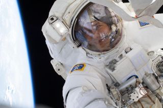 an astronaut in a white spacesuit floats outside the space station above Earth, his face visible through a clear glass dome over his face