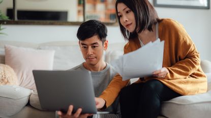 A man sits on the floor in front of the sofa where a woman sits as they look at a laptop and paperwork together.