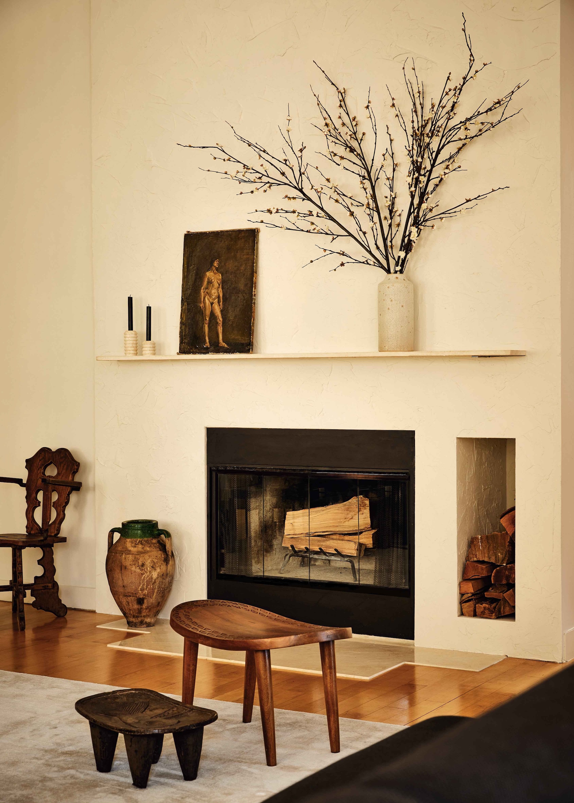 A white space by a modern fireplace with lots of naturals wood furniture pieces and a splay of twiggy branches in a vase on the mantelpiece.