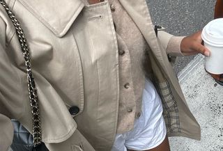 Claire Most wearing a tan trench coat, tan cardigan, and white shorts while walking and drinking a coffee.