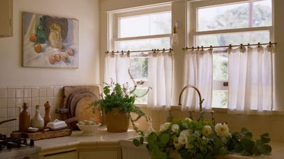 Sunlit rustic kitchen counter with cream tiles, potted herbs, fresh flowers in an apron sink, and a still-life painting.