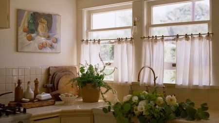Sunlit rustic kitchen counter with cream tiles, potted herbs, fresh flowers in an apron sink, and a still-life painting.