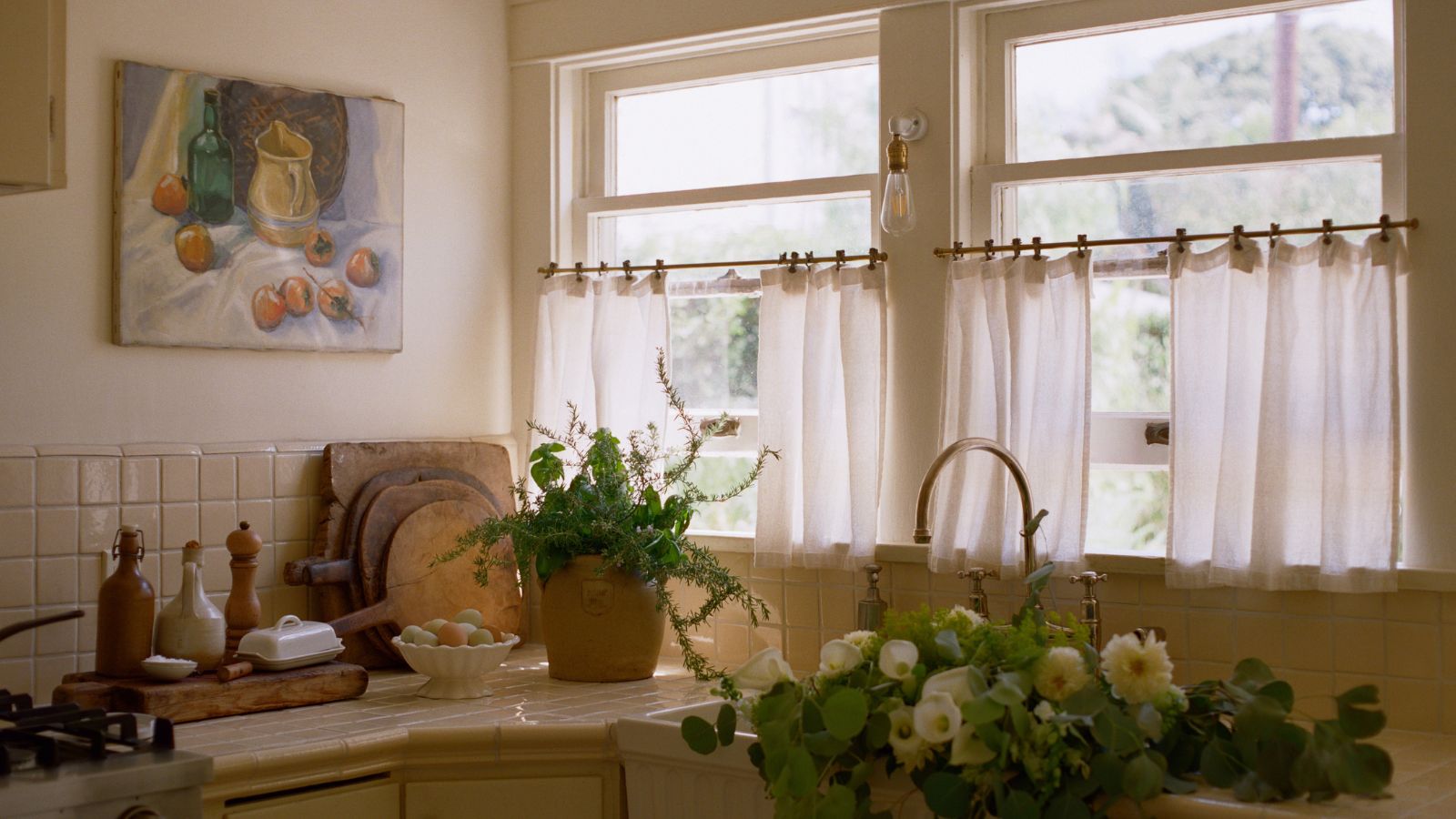 Sunlit rustic kitchen counter with cream tiles, potted herbs, fresh flowers in an apron sink, and a still-life painting.