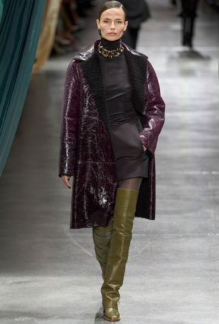 Model walking the Fendi fall runway show wearing a purple leather coat with a turtleneck, miniskirt, tights, and over-the-knee green boots.