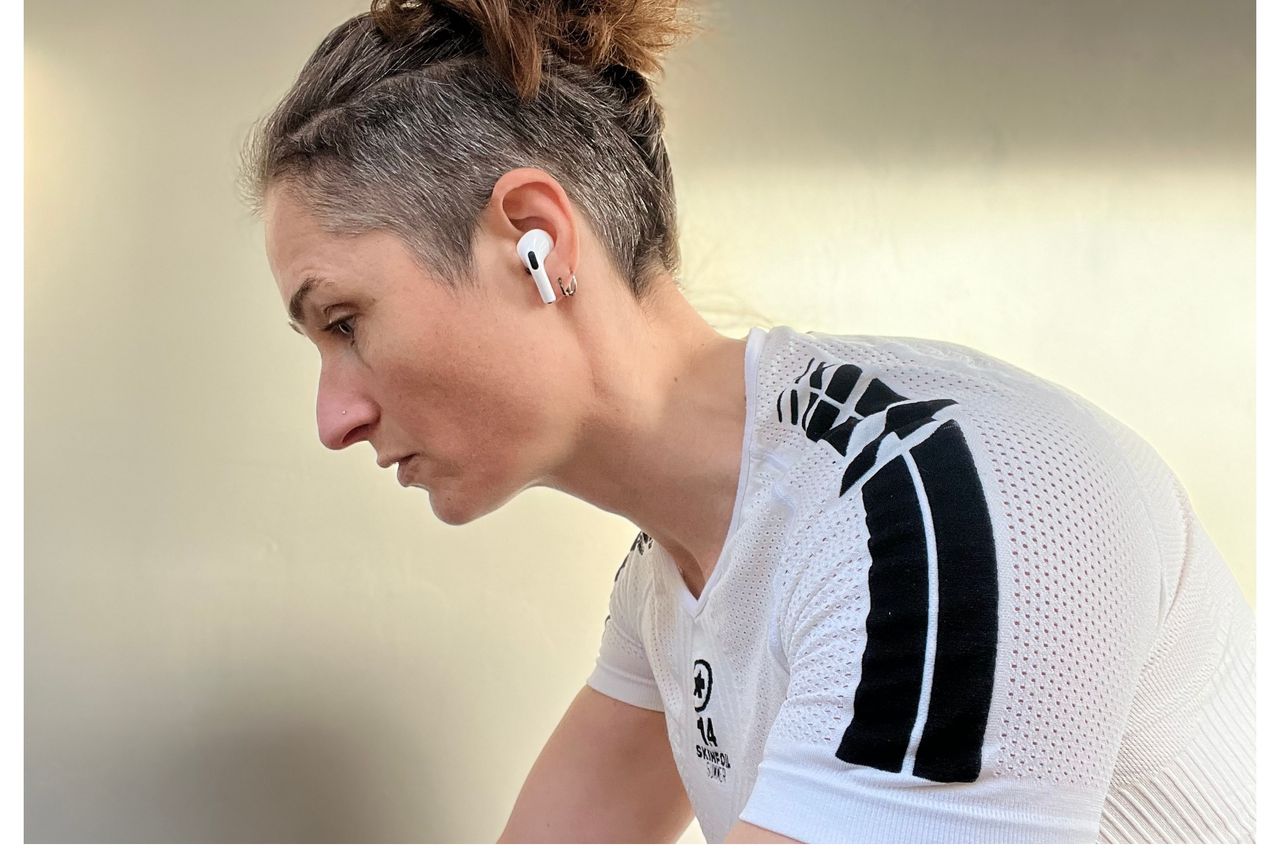 The best headphones for cycling are a pair of Apple Airpods Pro, as shown being worn in the ears of a woman side on with her face pointing to the left. In the image she has a white top on with a black stripe on the shoulder against a cream background