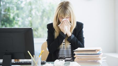 An office worker sitting at her desk at work blows her nose.