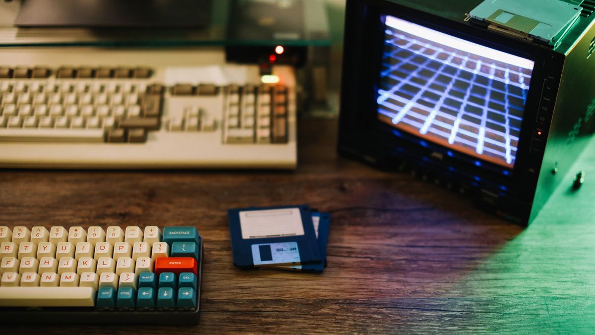 A wooden desk with a retro monitor, keyboard, and floppy disk sitting on top to represent legacy tech skills. Decorative: the desk is warmly-lit, with cold green light entering the frame on the right-hand side.