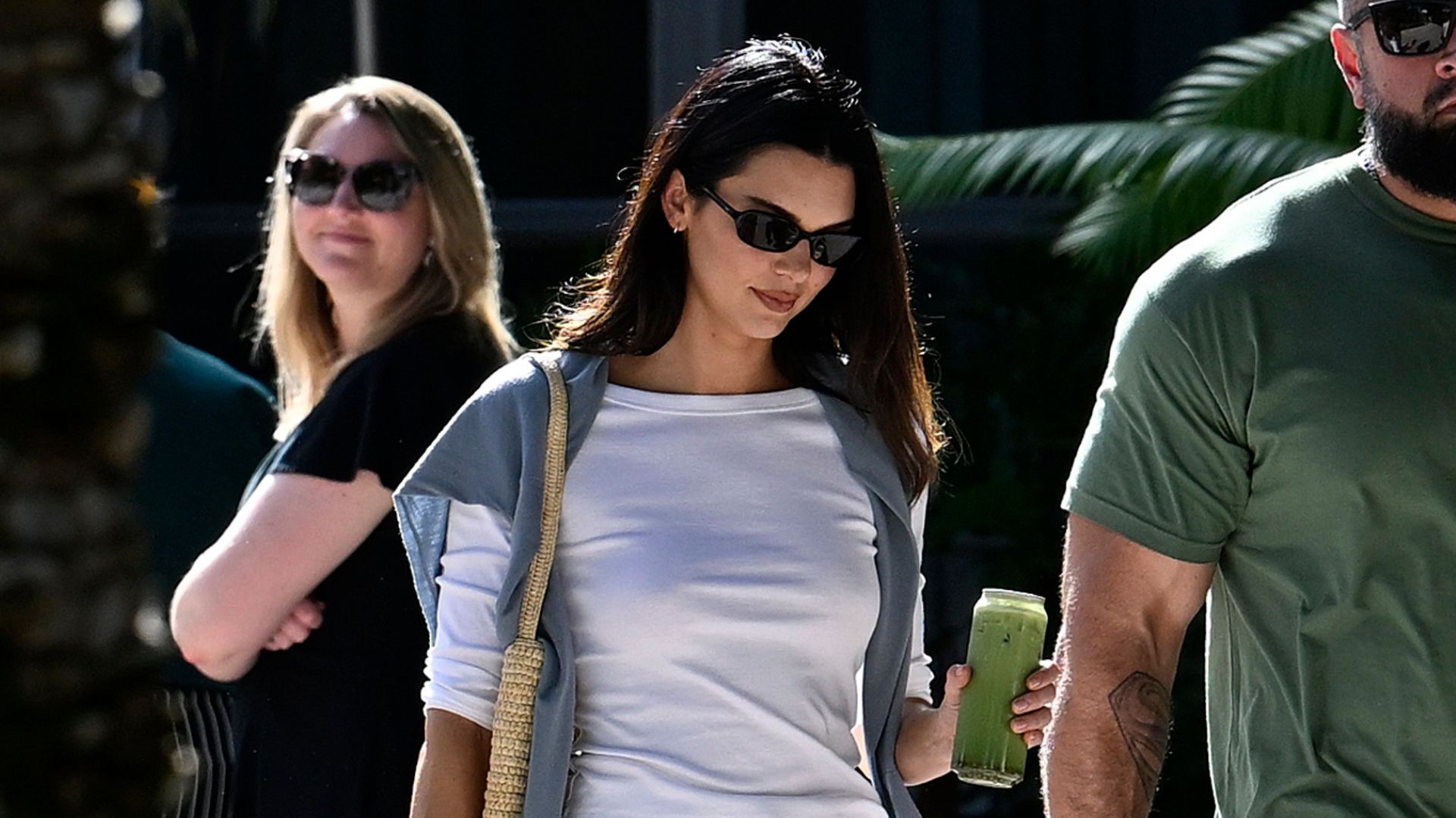 Kendall Jenner walks outside wearing white straight-leg trousers with a white boat-neck t-shirt and black slipper shoes.