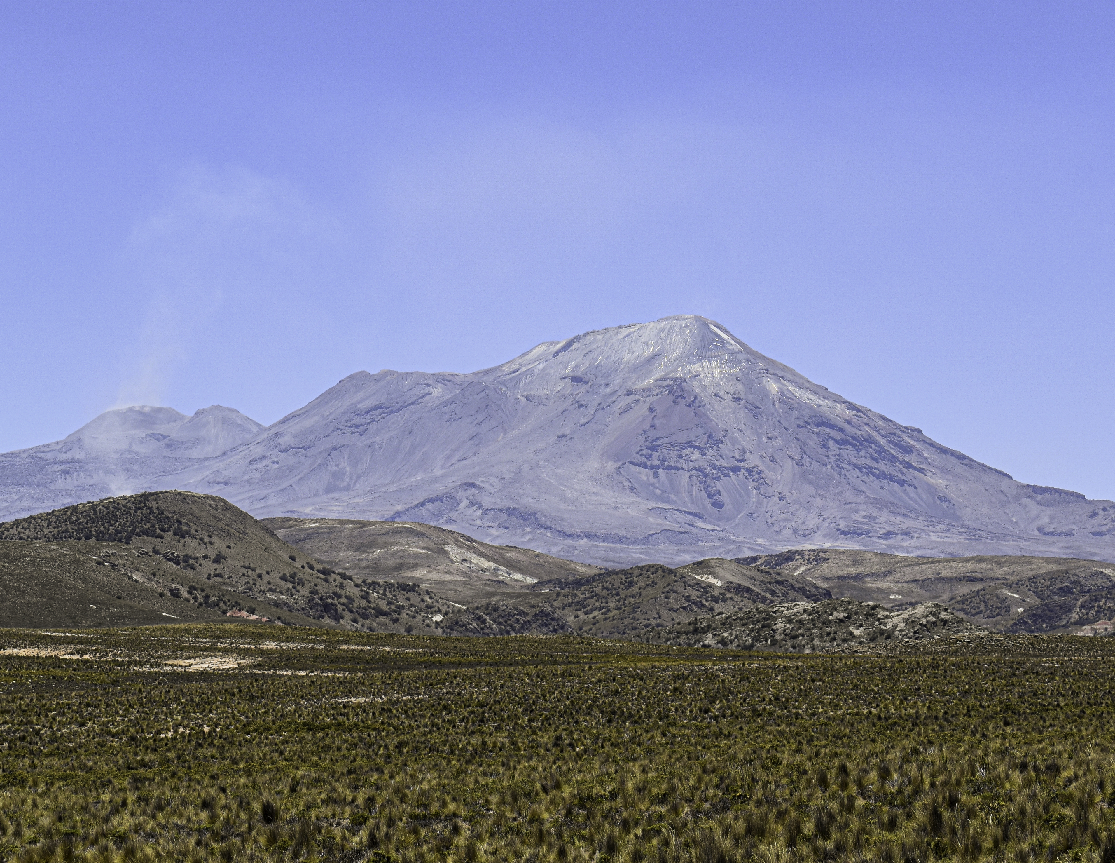 A series of brown peaks loom over green and brown hills and meadows in the foreground, all under a sunny blue sky.