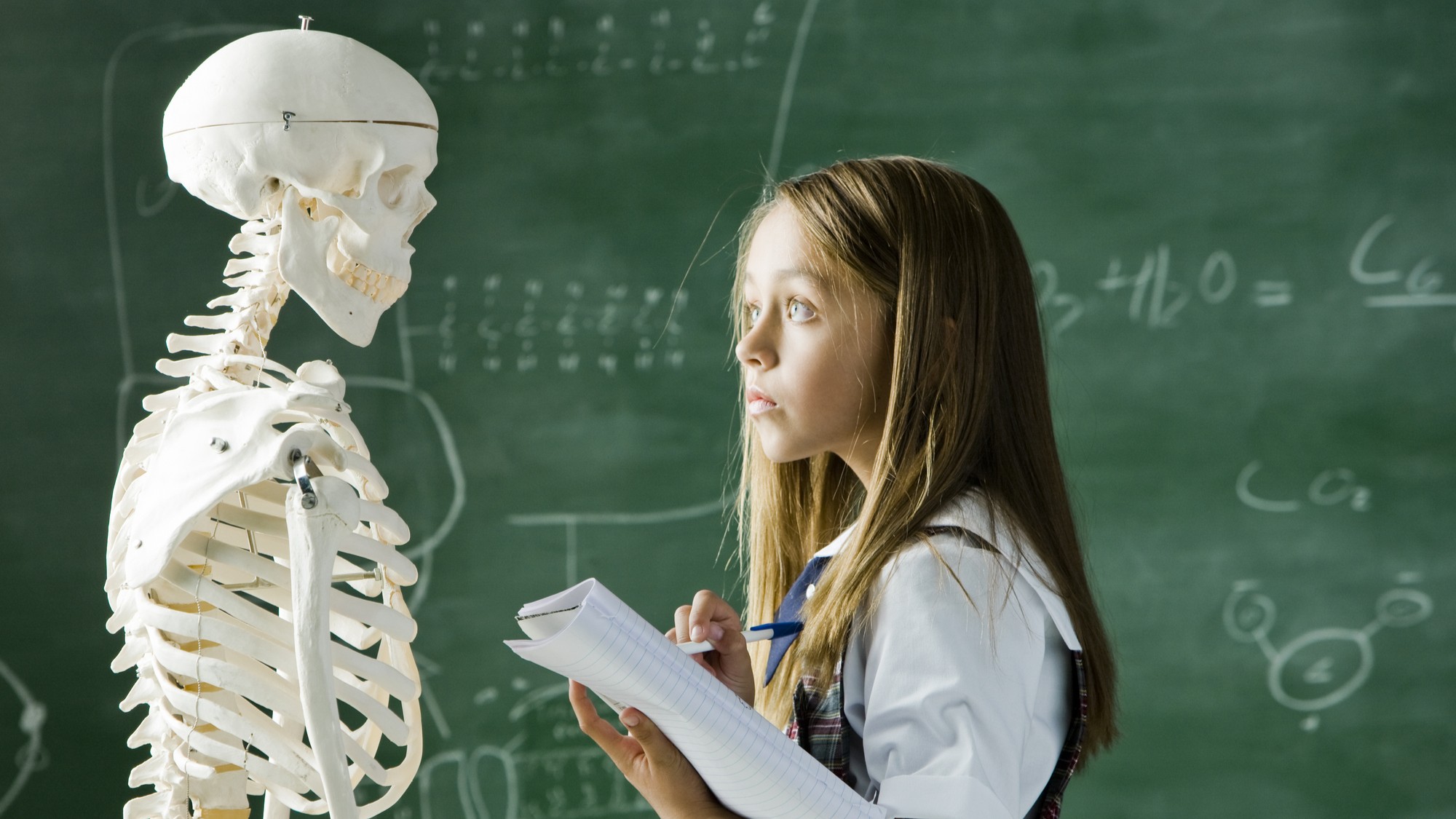 A girl in a uniform contemplates the model of a human skeleton. She is taking notes and is in science class.