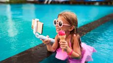 A young child influencer films herself with a smartphone while holding an ice cream cone in a swimming pool. 