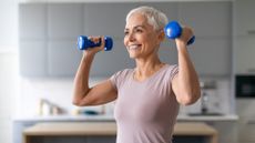 woman from waist up, holding light blue dumbbells in each hand ready to do an overhead press, smiling. she's in a kitchen setting that's blurred in the background.