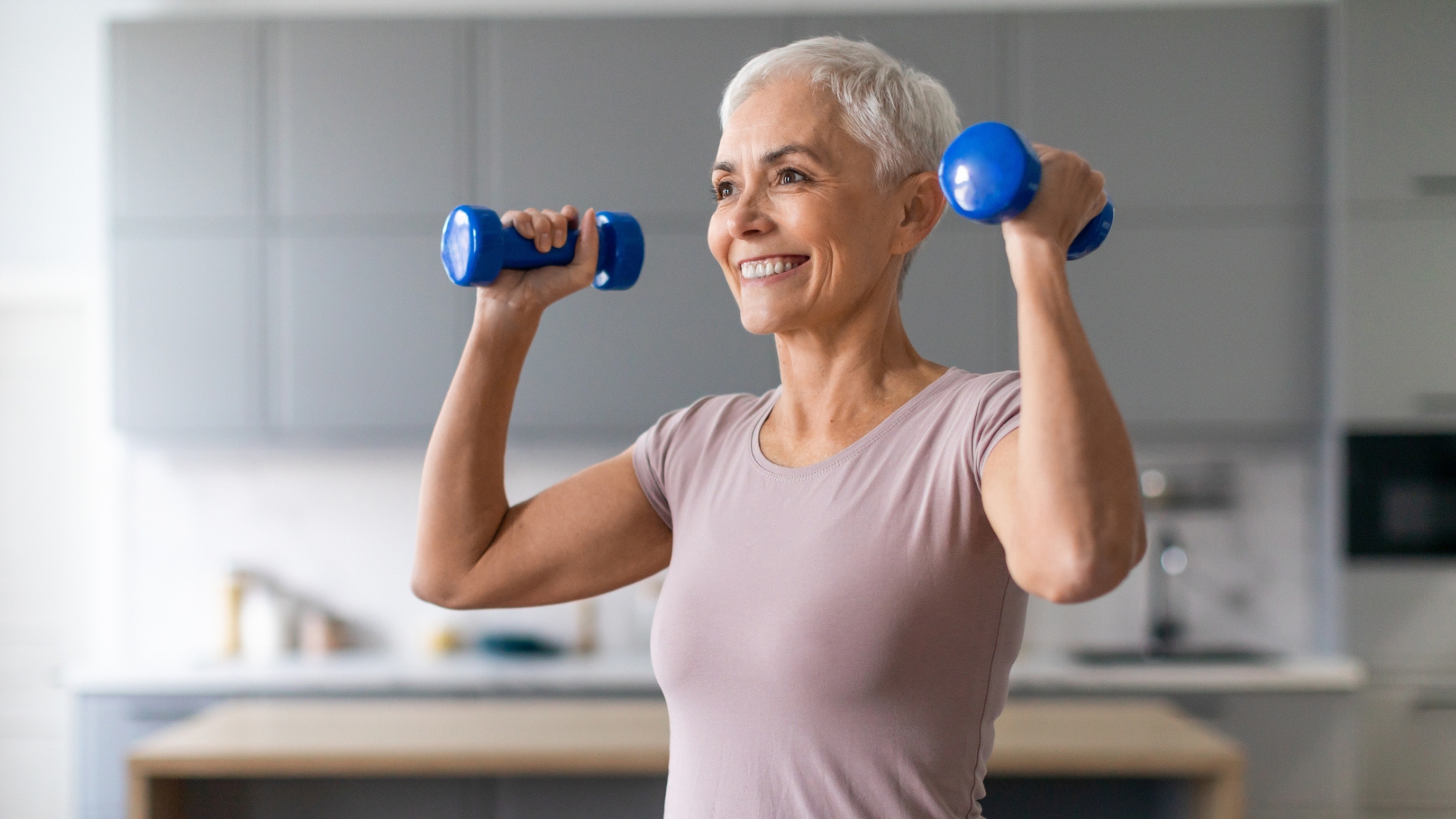woman from waist up, holding light blue dumbbells in each hand ready to do an overhead press, smiling. she&#039;s wearing a light pink tshirt in a kitchen setting that&#039;s blurred in the background. 