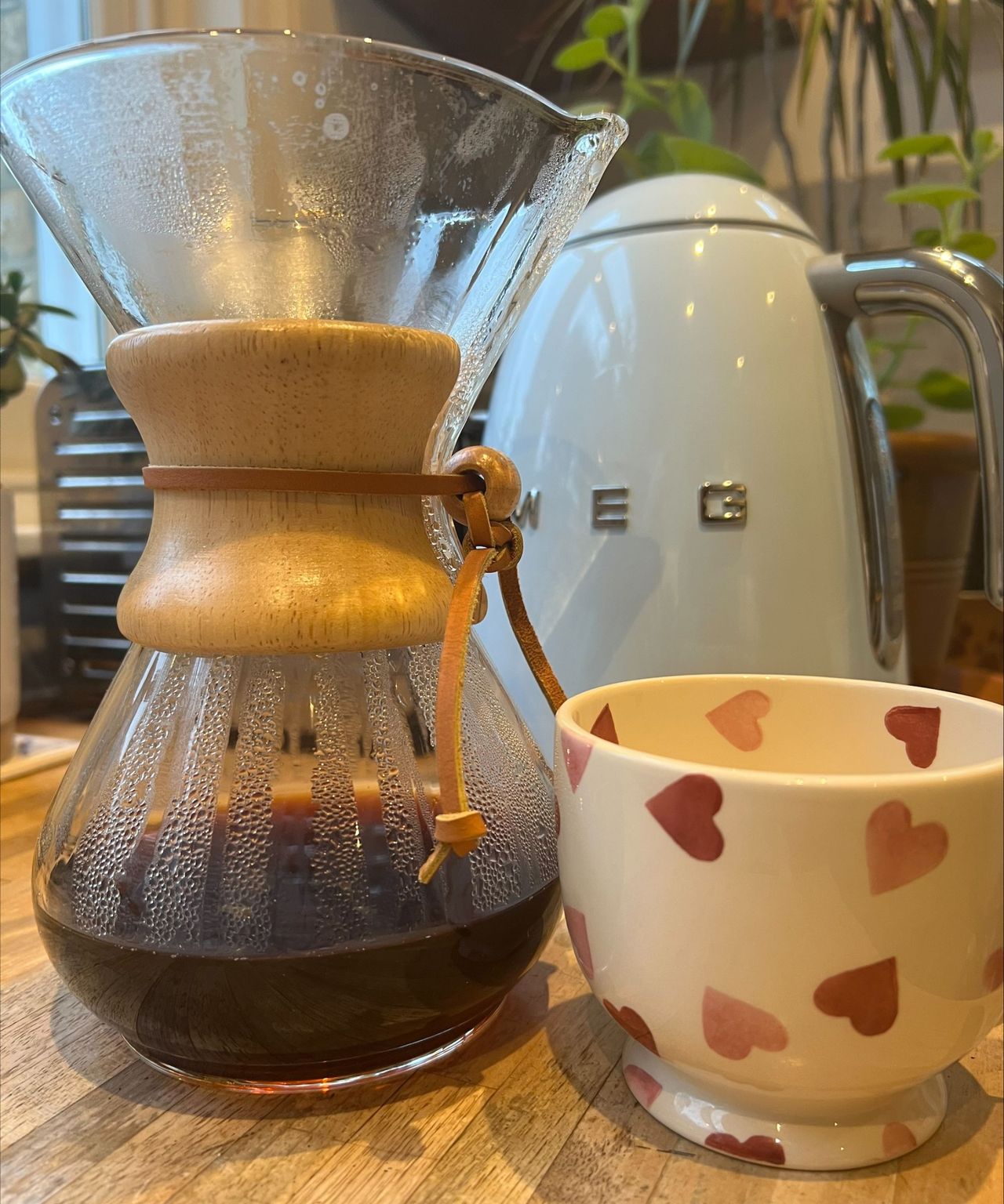 A CHEMEX coffee maker standing on a wooden countertop, filled with freshly brewed black coffee, beside a white coffee cup with pink hearts. Behind is the corner of a window, the side of a Dualit toaster, a baby blue SMEG kettle, and a houseplant.