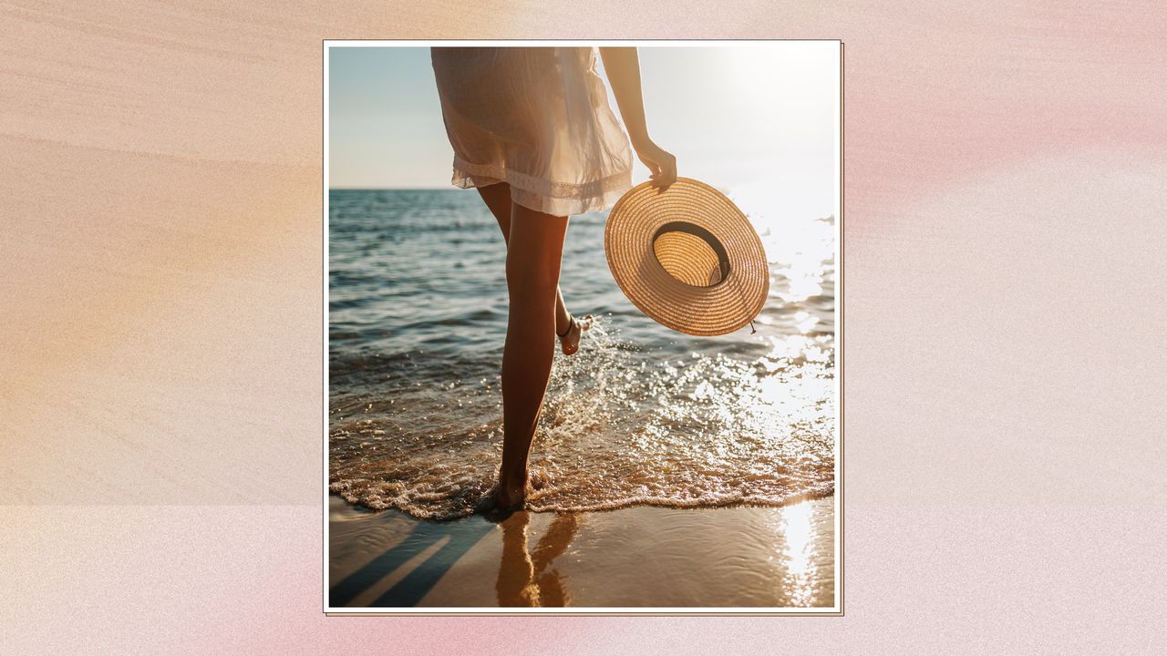 Back view of woman&#039;s legs walking into the sea as she carries a sunhat, in a white frame and on a pink watercolour-style background