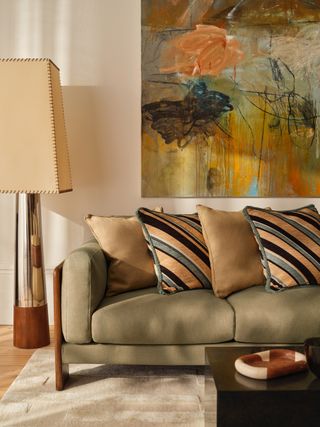 Image of a living room with a sage green sofa that has lots of beige and light blue throw pillows lined up on it. There is an abstract piece of art hanging above the sofa on a white wall. The floor lamp next to the sofa is chrome and wood with an oversized lamp shade that is cream with brown stitching.