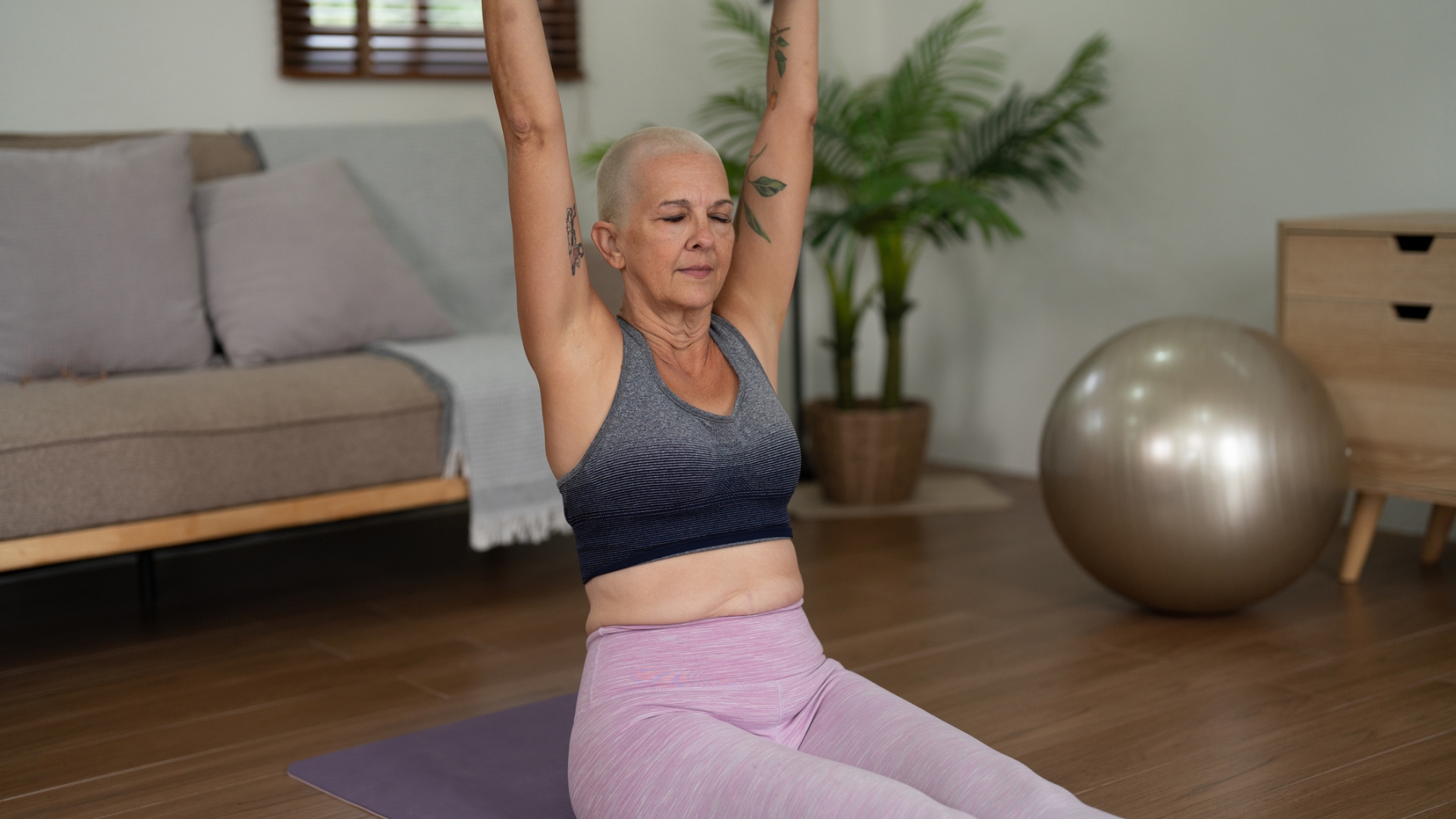 woman facing the camera sat on an exercise mat on wooden floor with her legs in front and arms stretched overhead. she's wearing light pink leggings and grey crop top. there's a sofa a golden balance ball behind her.