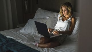 A woman with blonde hair works on her laptop in bed close to bedtime