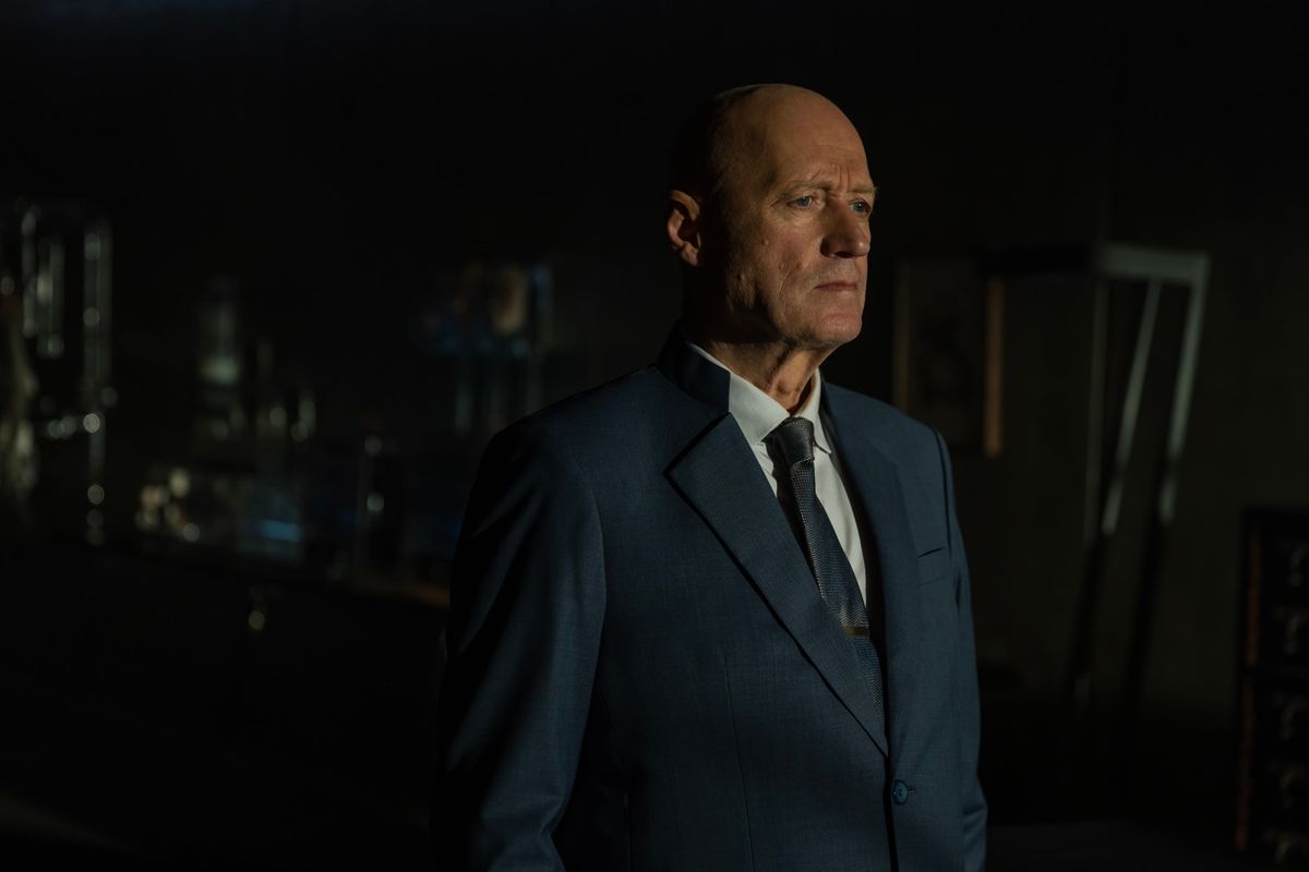 Still from the TV show Alien: Earth. A man wearing a suit is standing in a dark room, looking off into the distance.