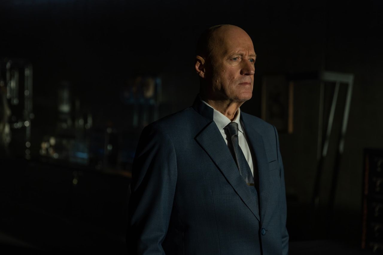 Still from the TV show Alien: Earth. A man wearing a suit is standing in a dark room, looking off into the distance.