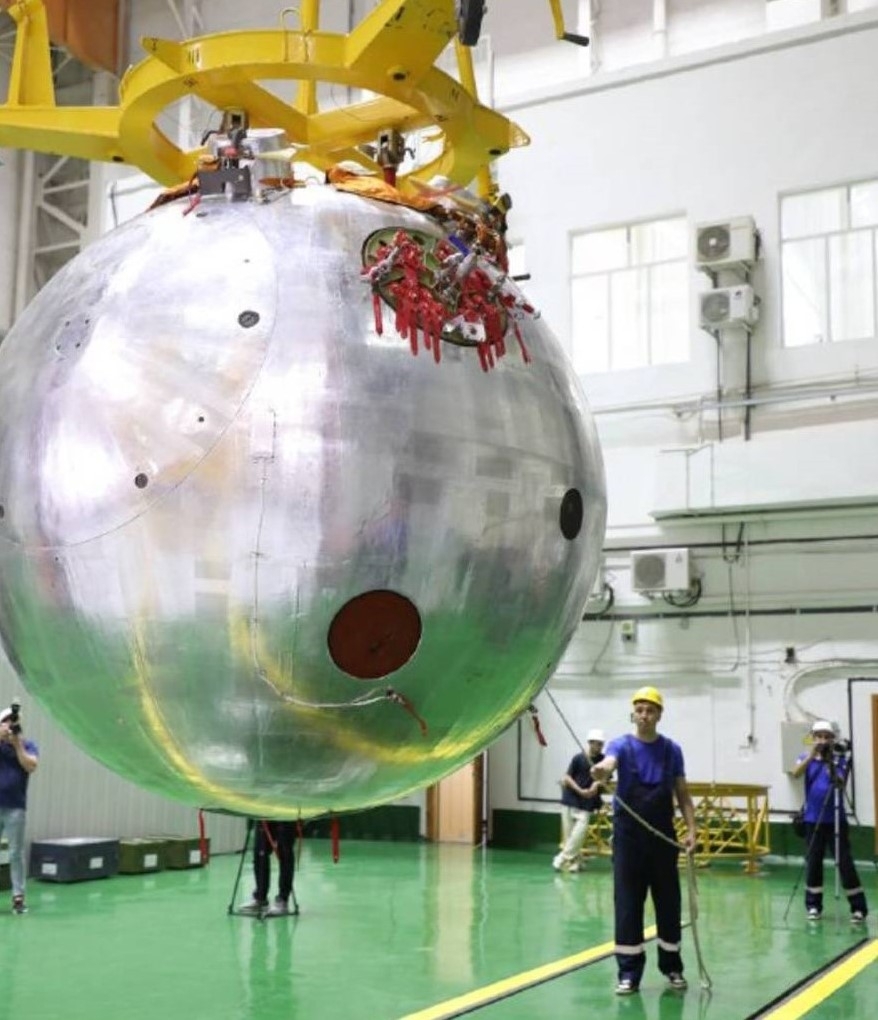 People wearing blue shirts, black pants and yellow hardhats work around a giant metal ball hung in the center of a room