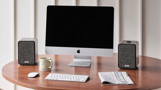 A pair of Ruark Audio MR1 Mk3 Bluetooth speakers on a wooden desk, sitting either side of a silver iMac computer. There is a keyboard, a mouse, a newspaper and a mug on the desk.