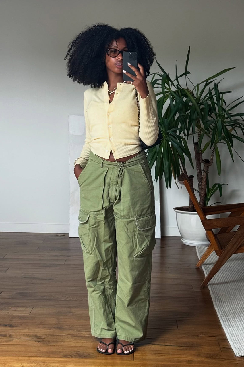 A woman wearing a yellow cardigan sweater, green cargo pants, black thong sandals and black frame sunglasses