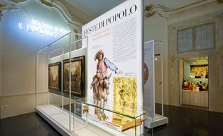 Black glossed floor, white walled room, with a gold frame detail to the the ceiling, white neon sign on the far left wall, floor standing exhibition piece in the centre of the room about Feste Di Popolo and artwork in a glass frame, door way looking out to a doll exhibition