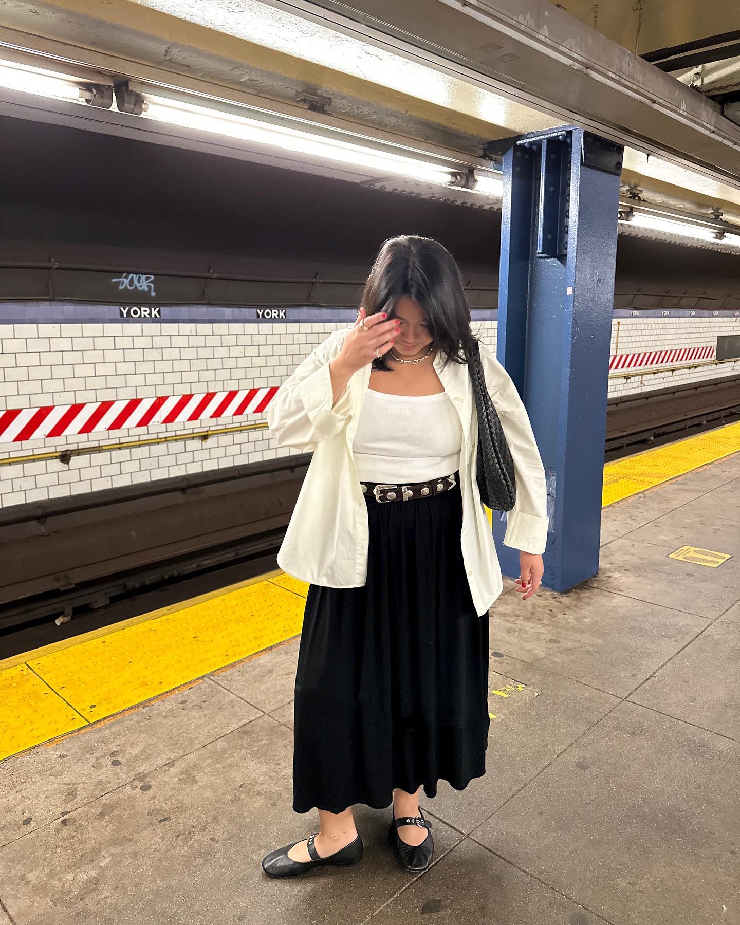 stylish mid-size woman poses on the subway platform in NYC wearing a white button-down shirt, white tank top, studded black belt, black full skirt, and black Mary-Jane flats