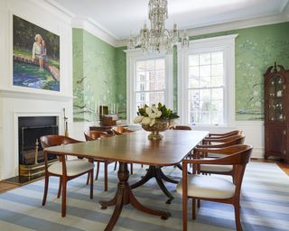 A dining room with green printed wall paper, traditional wooden furniture and white fireplace with family portrait hung above