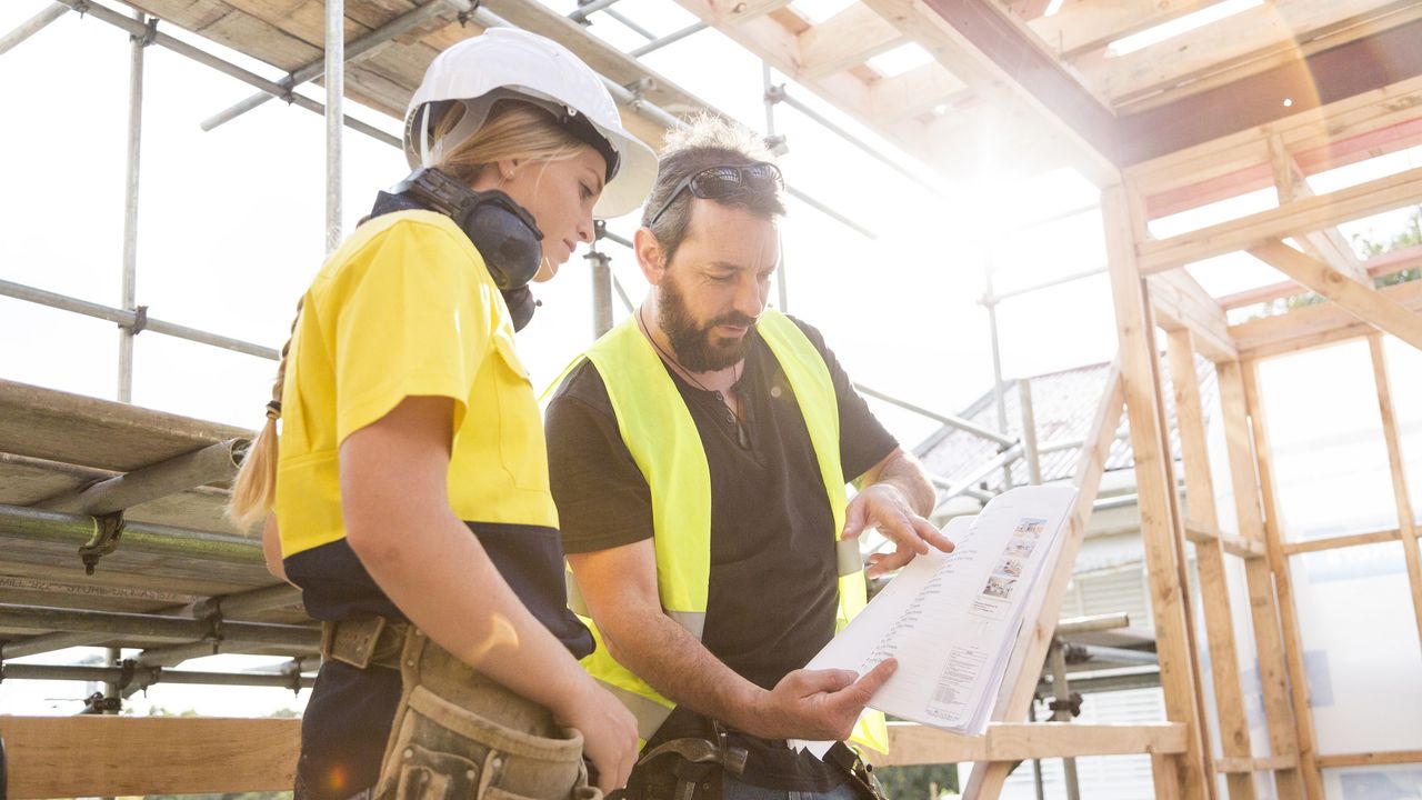 A woman and man wearing hard hats look at blueprints in a house that&#039;s being built.