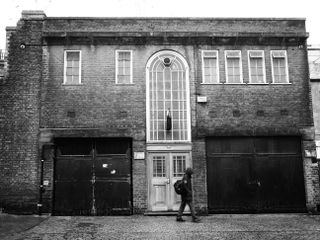 Mono image of a person walking past a building, taken on the OM System OM-3 with the OM System M.Zuiko 17mm f/1.8 II