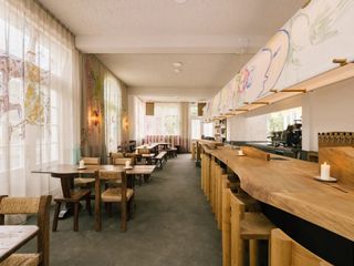 A lively, warmly welcoming bar features organically shaped wooden bar counters and matching modern rustic tables, flowy courtains, and handpainted murals floating above it all.